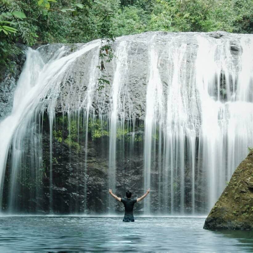Pria mengangkat tangannya ke atas sedang menikmati air terjun Damar
